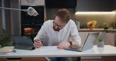Portrait of businessperson working with laptop and reading documents at home in kitchen. Man works with financial documents. Remote job and paperwork concept. Slow motion.