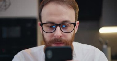 Close up of happy man working on mobile phone in kitchen at home at night. Attractive young male using smartphone chat typing browsing reflection eye glasses. Slow motion.