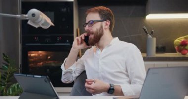 Cheerful man talking on a cell phone, laughing and working on a laptop. Home evening. Male freelancer using cellphone discussing business project with colleague. Kitchen on background. Slow motion.