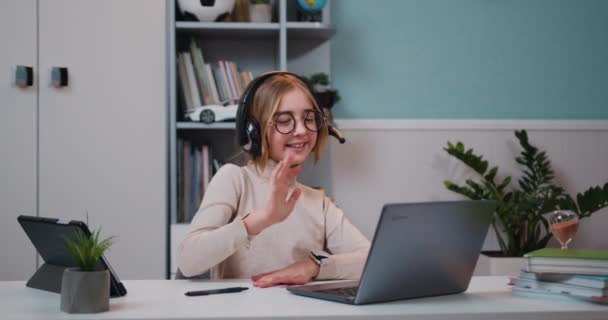 ヘッドフォンを装着したスマート白人の子供の女の子は 自宅のリビングルームでコンピュータ上の学習クラスを呼び出す社会的距離ビデオ会議の遠隔Web教師と話を手を振って スローモーション — ストック動画