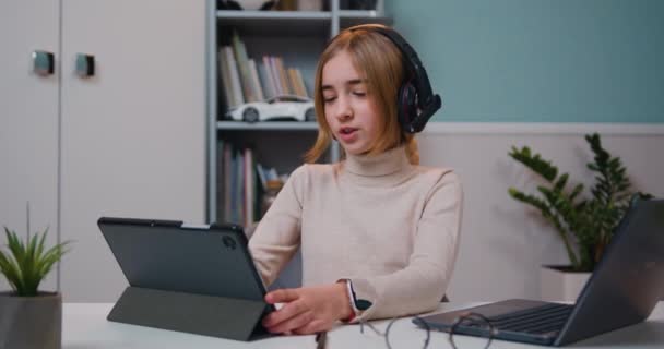 聪明漂亮的高加索10岁小女孩 戴着耳机 坐在客厅的桌子旁 看着平板电脑上的媒体 戴耳机的少女使用数码平板电脑和学习英语 — 图库视频影像
