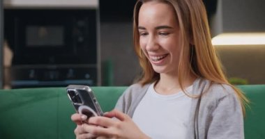 Kanepede oturup modern odada akıllı telefon kullanan neşeli genç bir kadın. Cep telefonuyla konuşurken gülümseyen ve düşünen güzel bir kız. Yavaş çekim.