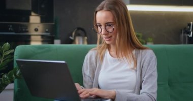 Young woman in glasses sits at on sofa at laptop computer working at home. Female freelancer or student listens to an online conference in the kitchen. Slow motion.
