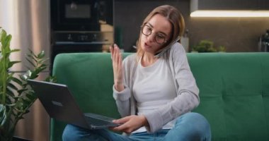 Evde dizüstü bilgisayarla çalışan ve modern mutfaktaki kanepede oturan akıllı telefonla konuşan güzel bir genç kadın. Bayan müşteri online alışveriş siparişi teslimatını onaylamak için cep telefonu görüşmesi yapıyor. Yavaş çekim.