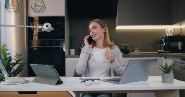 Genç, mutlu bir kadın elinde kahve fincanıyla akıllı telefonuyla bir telefon görüşmesi yapıyor. Modern mutfakta gülümseyen serbest çalışan kadın telefonda uzaktan konuşuyor. Yavaş çekim.