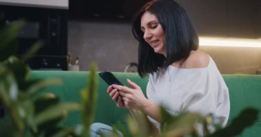 Çekici bir kadının portresi kanepede oturuyor ve evdeki modern mutfağın arka planındaki akıllı telefondan mesaj yolluyor. Ev mutfağında cep telefonu kullanan düşünceli genç bir kadın. Yavaş çekim