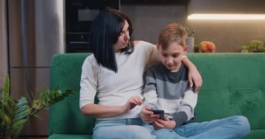 Mutfağın arka planında kanepede otururken annesiyle birlikte akıllı telefonda oyun oynayan 9 yaşındaki yakışıklı beyaz çocuk. Cihazlar, genç nesil internet teknolojisi konsepti. Yavaş