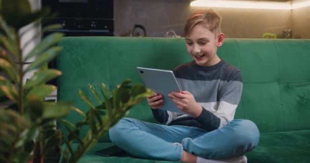 Uśmiechnięty Chłopiec Siedzący Kanapie Skrzyżowanymi Nogami Trzymający Tablet Sms Ujący — Wideo stockowe