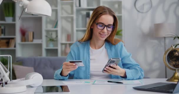 オンラインストアで買い物に銀行カードを使用している女性 部屋のテーブルの上にスマートフォンを介してオンラインで支払うためにクレジットカードコードを入力する若い女の子 スローモーション — ストック動画