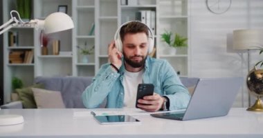 Modern, mutlu ve rahat kulaklıklı bir adam elinde cep telefonuyla video izliyor ve favori müzik podcast 'ini ya da internet sosyal medyasını dinliyor. Yavaş çekim.