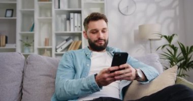 Çekici bir gencin portresi kanepeye oturmuş modern bir dairede telefon tebessümü kullanarak akıllı telefon sosyal medya uygulamalarındaki mesaj içeriğini internetteki kayıt teknolojisine yolluyor. Yavaş