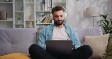 İş adamı kablosuz kulaklıklar evdeki yaratıcı genç adamın dizüstü bilgisayarını kullanarak koltukta oturur ve interneti tarar ve sosyal medyayı yönetir. E-hocayla öğren, bilgiyi geliştir video kullan, modern teknoloji