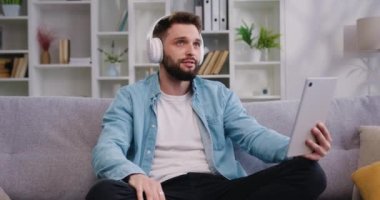 Sakallı adam video görüşmesiyle konuşan kulaklık takıyor. Video çağrı uygulaması kullanarak internetteki iletişimde yer alıyor. Erkek serbest çalışan evde dijital tablet üzerinde sanal toplantı yapıyor. Yavaş çekim.