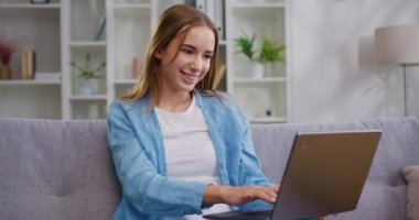 Çekici, mutlu, kaygısız genç kız evdeki kanepede dinleniyor ve dizüstü bilgisayarında arkadaşından gelen mesajı okuyor. Uzaktan çalışma konsepti. Yavaş çekim.