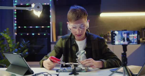 男の子Vloggerは スマートフォンを使用してオンラインストリーミングを行う彼の科学プロジェクトで電子機器やハンダ付けワイヤや回路基板を研究しており 自宅で信者を教えています スローモーション — ストック動画