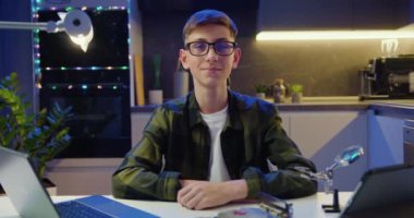 Genç bir çocuğun portresi. Masasında dizüstü bilgisayarla oturmuş gece mutfaklarındaki kameralara bakıyor. Yavaş çekim.