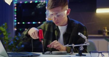 Koruyucu gözlüklü yakışıklı meraklı genç akşamları evdeki elektronik panelde lehimliyor. Yavaş çekim.