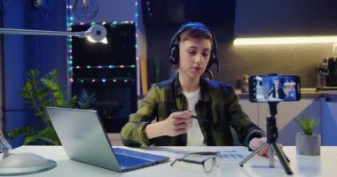 Ergen çocuk, öğretmenini ve sınıf arkadaşlarını cep telefonuyla karşılıyor. Üç ayaklı cep telefonuyla evdeki akşam odasında oturuyor. Erkek, modern sosyal medya vlog uygulamasında sanal video görüşmesi yapıyor. Yavaş çekim.