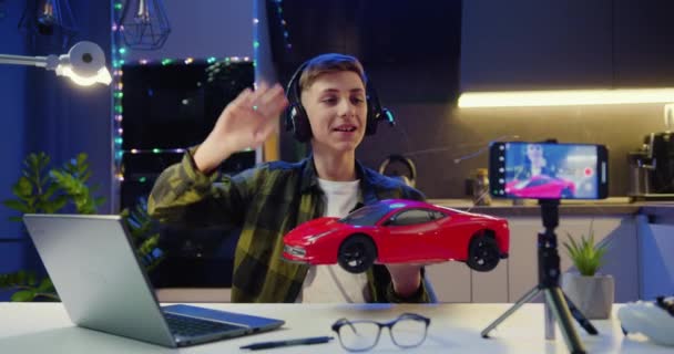 インターネットの聴覚のためのスマートフォンでビデオを記録し 新しいおもちゃの車を示すよい笑顔の十代の男の子ブロガー スローモーション — ストック動画