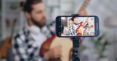 Ekran akıllı telefonunu kapat. Genç erkek blogcu elektronik gitar teli çalan kulaklık takıyor ve müzik videosu kaydediyor. Online eğitim öğrenme konsepti öğretiyorum. Yavaş çekim.