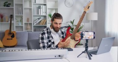 Yetenekli bir sanatçı akıllı telefonuyla video kaydediyor. Oturma odasında oturmuş gitar çalıyor. Erkek müzisyen vlog için cep telefonuyla video çekiyor. Yavaş çekim.