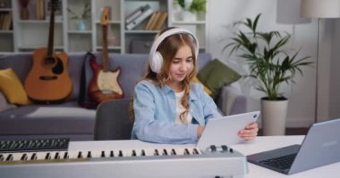 Kulaklık takan neşeli kız ev müziği stüdyosunda müzik dinliyor. Güzel mutlu çocuk, rahat oturma odasında dijital tablet kullanıyor dans ediyor ve şarkıdan keyif alıyor. Yavaş çekim.