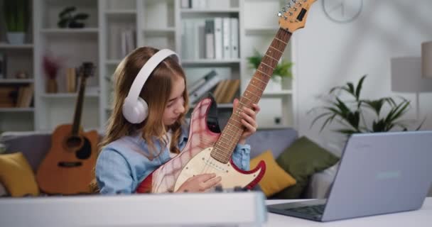 年轻的女音乐家带着耳机在笔记本电脑监视器前使用在线视频课程来提高技巧 家庭吉他手正在家里练习曲调 慢动作 — 图库视频影像