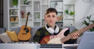 Fotoğraftaki gülümseyen çocuk kameraya bakıyor. Avrupalı erkek, ev stüdyosunda dizüstü bilgisayarla elektro gitar öğreniyor. E-öğrenme, online eğitim konsepti. Yavaş çekim.