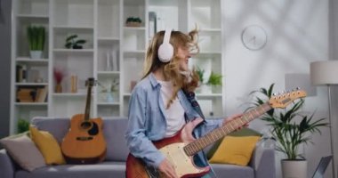 Uzaktan kumandalı beyaz kulaklık takan neşeli genç kız evde elektro gitar çalıyor ve modern müzikten zevk alıyor. Müzik aşığı konsepti. Yavaş çekim.