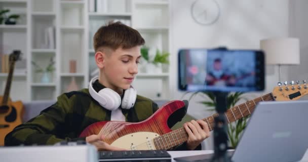 10代の男の子がスマートフォンにギターレコードビデオを再生します ブロガーの男は 初心者 インターネット聴覚のための文字列楽器クラスのためのチュートリアルレッスンを作成します スローモーション — ストック動画