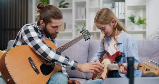 漂亮的高加索10岁小女孩正在练习弹电吉他 而她的父亲在教她 在音乐课上花时间和她在一起 同时坐在沙发上 慢动作 — 图库视频影像