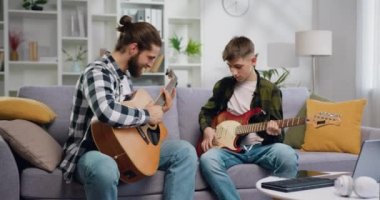 Baba ve oğul evde gitar çalar. Babası çocuklara müzik aletleri çalmayı öğretiyor. Birlikte mutlu vakit geçirmek. İnsanlar ve müzik konsepti. Yavaş çekim.