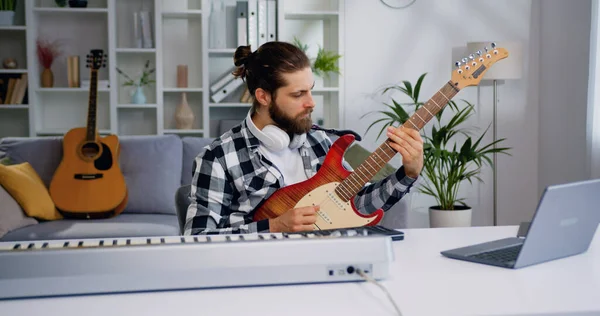 Attaractive adam evinin oturma odasında dizüstü bilgisayarıyla müzik kaydediyor. Erkek, evde dizüstü bilgisayar kullanarak gitar çalıp şarkı söylüyor. Hobi, müzik ve gençlik kavramı.