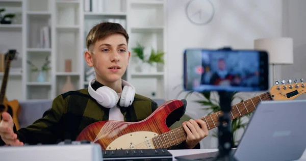 10代の男の子がスマートフォンにギターレコードビデオを再生します ブロガーの男は 初心者のためのチュートリアルレッスンを作成します インターネット聴覚のための文字列楽器クラス — ストック写真