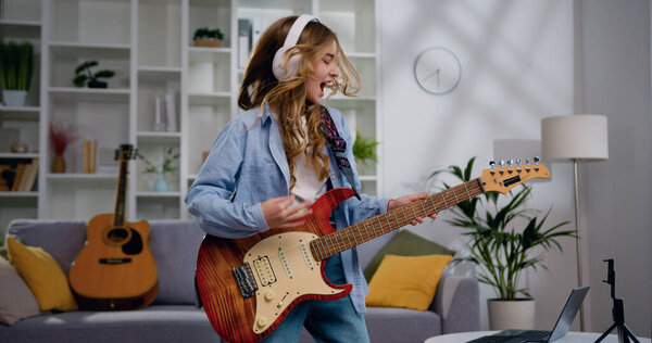 Joyful teen girl wearing white headphone singing in remote control playing electric guitar dancing at home enjoying modern music. Music lover concept.