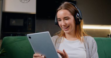 Mutlu gülümseyen kadın dijital tablet taraması yapıyor, internetten kulaklıkla video izliyor. Kanepede mobil cihazlı mutlu kız sosyal iletişim ağı.