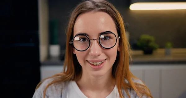 Yakından güzel bir kız kameraya bakıp gülümsüyor ve evdeki modern mutfakta gözlüğünü çıkarıyor..