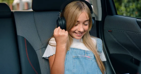 stock image Beautiful positive carefree 12-year-old blond girl in trendy clothes wears wireless earphones searching for something on tablet gadget and reacts emotionally after finding while sitting in autos