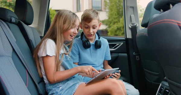 stock image Attractive joyful blond 10-12-year-old kids sitting on cars backseat and enjoying games on tablet gadget,entertainment during car trip