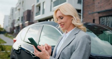 Çekici ve zeki yetişkin iş kadını, elinde akıllı bir telefonla, yerleşim yerindeki otoparkta siyah bir arabanın yanında duruyor. Açık havada. Yoğun yaşam tarzı