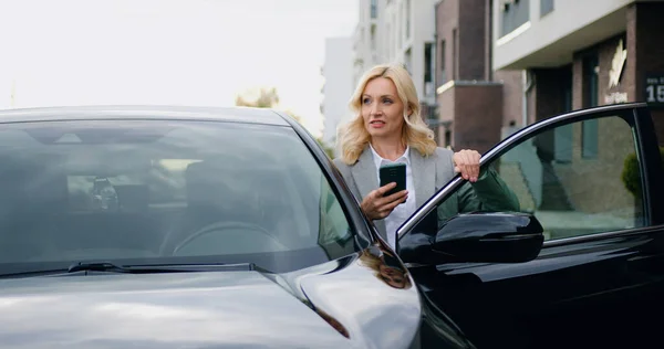 stock image Good-looking positive and successful business 40-aged woman in formal clothes typing texting on shes mobile,standing near auto in outdoors on parking