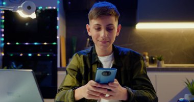 Gülümseyen mutlu genç çocuk akıllı telefon uygulamaları kullanıyor, oyun oynuyor, sosyal medyada sohbet ediyor, akşam evde sörf yapıyor..