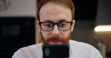 Geceleri mutfaktaki cep telefonuyla çalışan mutlu adama yaklaş. Akıllı telefon sohbeti yapan çekici genç bir erkek. Gözlük takıyor..