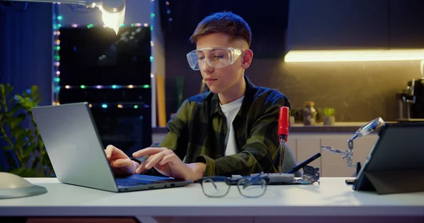 晚上在家 在笔记本电脑上修改培训视频时 聪明的少年学生戴着防护眼镜 用焊铁焊接面板 工程师焊接微电路 — 图库照片