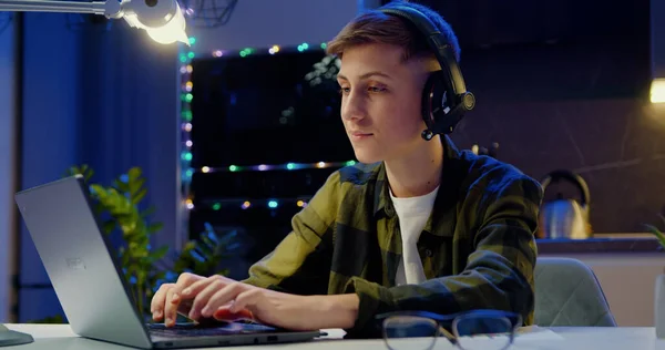 深夜时分 身穿无线耳机在笔记本电脑前为学校的一个项目工作的男孩微笑着 男孩在键盘上输入短信 孩子们在网上消磨时间 — 图库照片