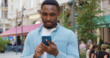 Gündüz vakti sokak kafesinin yanında cep telefonuyla günlük kıyafetlerini giyen çekici, Afro-Amerikan bir genç.