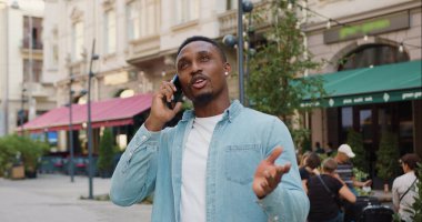 Güzel görünüşlü, gülümseyen, sıradan giysiler içindeki genç, siyah tenli adamın ön görüntüsü. Açık havada, sokak kafesi yakınlarında yaptığı telefon konuşmasından keyif alıyor.