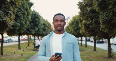 Çekici, gülümseyen, güler yüzlü, yeşil ağaçlı ve güneşli bir günde kameraya bakan, günlük kıyafetlerle gezen genç, siyah tenli bir adamın önü.