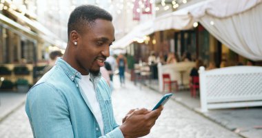 Yakışıklı, neşeli, genç, siyah tenli, stil sahibi bir adam açık hava kafeleri ile şehir caddesinde duruyor ve akıllı telefonunu, ön görüntüsünü kullanıyor.