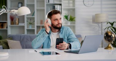 Modern, mutlu ve rahat kulaklıklı bir adam elinde cep telefonuyla video izliyor ve favori müzik podcastını veya internet sosyal medyasını dinliyor..